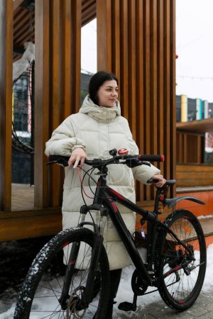 Eine Frau steht neben ihrem Fahrrad in einer verschneiten Landschaft. Sie ist warm angezogen für das kalte Wetter. Das Fahrrad steht auf dem schneebedeckten Boden.
