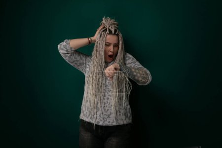 A woman with dreadlocks is standing upright in front of a solid green wall. She appears confident and relaxed, with her hair falling freely around her shoulders. The green wall provides a simple