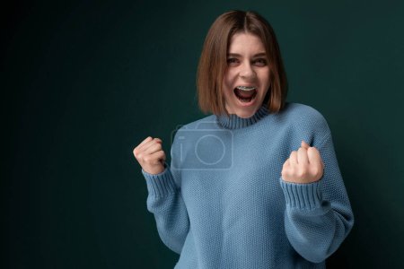 A woman wearing a blue sweater is shown in the act of making a fist gesture, conveying determination or anger. Her hand is clenched tightly with fingers folded inwards, symbolizing strength or resolve