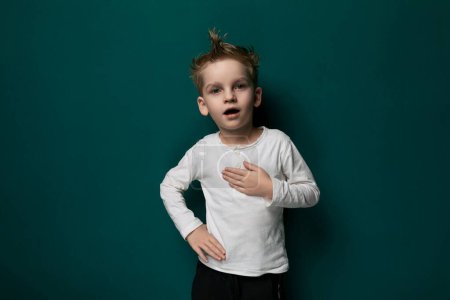 A young boy is standing in front of a solid green wall, looking straight ahead. He appears calm and composed, wearing casual attire. The wall provides a simple background, emphasizing the boys