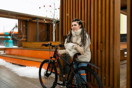 A woman with dreadlocks is seated on a bicycle, appearing relaxed and confident. She is wearing casual clothing, and the bike is stationary in an urban setting. The focus is on her unique hair and the