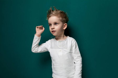 A young boy is standing upright against a bright green wall, his hands hanging loosely by his sides. The background is simple and unobtrusive, showcasing the boys posture and expression.