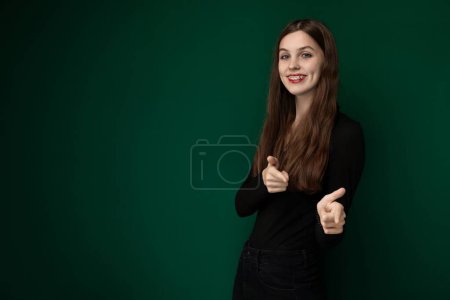 Una mujer con un gesto de mano que muestra un signo de pulgares hacia arriba se para frente a un fondo verde sólido. Ella aparece alegre y positiva en su lenguaje corporal.