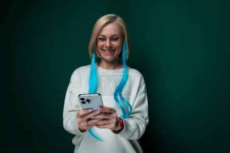 Una mujer con el pelo azul vibrante se ve sosteniendo un teléfono celular en su mano. Parece estar mirando la pantalla atentamente, posiblemente enviando mensajes de texto o navegando. El fondo está borroso, centrándose en la