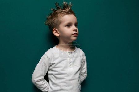 Un niño está de pie frente a una pared verde brillante, mirando curioso y ligeramente tímido. Él parece estar usando ropa casual y está sosteniendo un pequeño juguete en su mano.