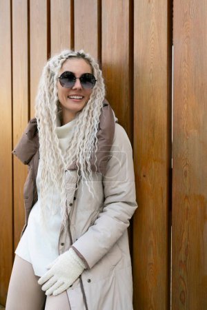 Une femme aux longs cheveux blancs est vêtue d'un manteau et de gants. Elle se tient debout dans une pose neutre, face à la caméra. Ses cheveux coulent autour de ses épaules, contrastant avec le manteau sombre. Les gants
