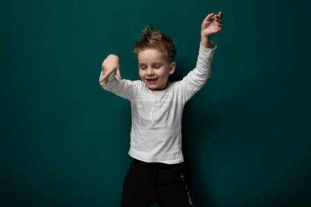 A young boy is standing upright in front of a vibrant green wall. He appears curious and engaged, looking straight ahead. The contrast between his clothing and the wall creates a striking visual.