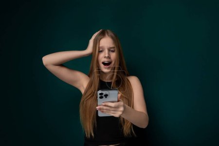 A woman is holding a cell phone in front of her face, appearing to take a selfie or video. She is looking directly at the phones screen with a neutral expression. The background is blurred out