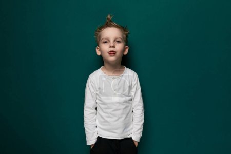 Ein kleiner Junge mit herausgestreckter Zunge steht vor einer leuchtend grünen Wand und zeigt einen verspielten Ausdruck. Er scheint Spaß zu haben und sich von seiner frechen Seite zu zeigen.