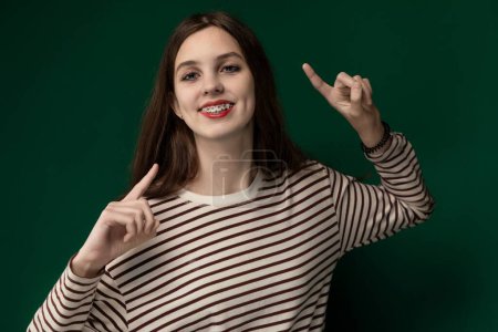 Une femme portant une chemise rayée étend ses doigts dans un geste de paix. Elle se tient debout sur un fond neutre, regardant directement la caméra avec une expression neutre sur son visage.