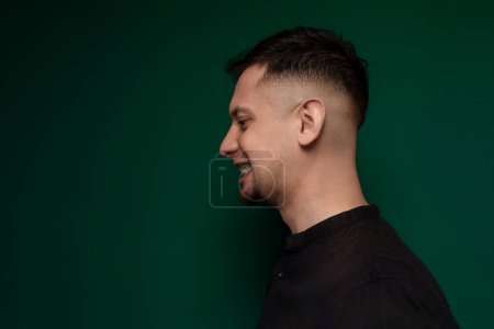 A man with a shaved head is standing with his back against a vivid green wall. He appears to be facing forward, with a calm expression on his face. The simplicity of the scene brings attention to the