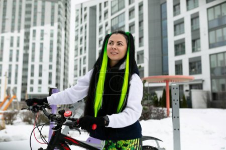 Eine Frau mit leuchtend grünen Haaren steht in urbaner Umgebung neben einem Fahrrad. Sie wirkt selbstbewusst und stilvoll und präsentiert ihre einzigartige Haarfarbe. Das Fahrrad bringt Farbe in die Szene