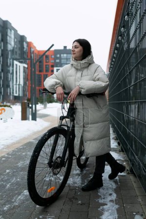 A woman is standing next to a bike in a snowy landscape. She is wearing winter clothing and appears to be preparing to ride the bike through the snow-covered terrain.
