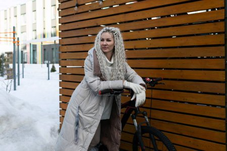 Eine Frau steht neben einem Fahrrad auf schneebedecktem Boden. Sie trägt Winterkleidung und blickt nach vorn. Das Fahrrad steht in einer verschneiten Landschaft.