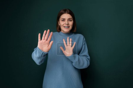 A woman wearing a blue sweater is standing with her hands raised up. She appears to be in a pose of surrender or excitement, with a neutral expression on her face.