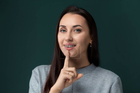 Eine Frau hält einen Finger an ihre Lippen und signalisiert Stille oder Stille. Sie scheint in einer Geste, die gemeinhin mit Geheimhaltung oder Geheimhaltung assoziiert wird, um Diskretion oder Geheimhaltung zu bitten.
