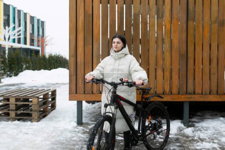 A woman is standing next to a bicycle in a snowy landscape. She is wearing winter clothing and appears to be examining the bike.
