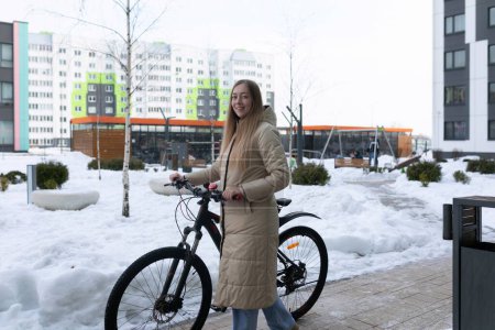 A woman dressed in winter clothing stands next to a bicycle covered in snow. The scene depicts snowy weather conditions with the woman appearing to assess or prepare the bike for use.