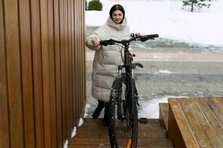 A woman is standing next to a bicycle in a snowy landscape. She appears to be dressed warmly for the cold weather, with snow covering the ground around her.