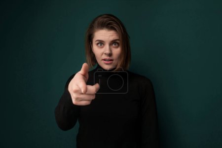 A woman is standing in front of a bright green background, pointing directly at the camera with a focused expression. She appears confident and assertive in her gesture towards the viewer.
