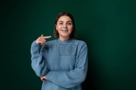 A woman wearing a blue sweater is pointing at an unseen object with a serious expression, indicating curiosity or interest. She is standing against a neutral background, focusing all attention on her