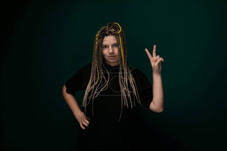 A woman with dreadlocks is extending her hand to make a peace sign gesture. She is dressed in casual clothing and is standing against a plain background.