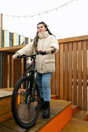 Una mujer está de pie junto a una bicicleta en una plataforma de madera. Ella está usando ropa casual y mirando hacia la distancia. La moto está aparcada junto a ella, y la escena parece estar en un tranquilo al aire libre
