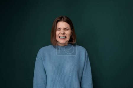 Eine Frau im blauen Pullover verzerrt ihren Gesichtsausdruck und zeigt Emotionen oder Reaktionen. Der Hintergrund ist einfach und konzentriert sich auf die Themen Aktionen.