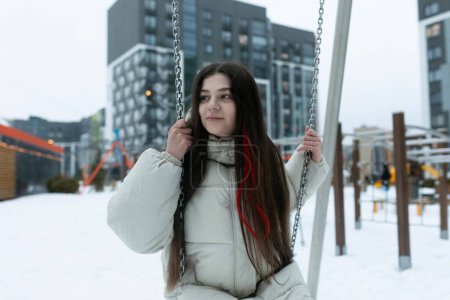 A woman is seated on a swing in a snowy landscape. She is bundled up in winter attire, with snow covering the ground around her. The swing is gently swaying back and forth as she enjoys the peaceful