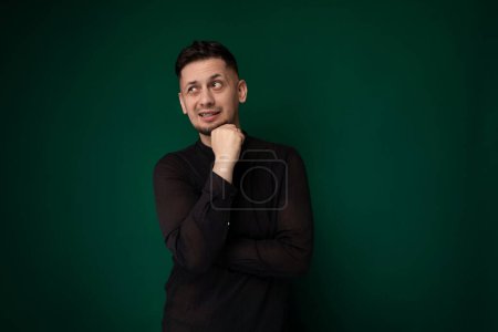 Ein Mann im schwarzen Hemd posiert für ein Foto und blickt mit selbstbewusstem Gesichtsausdruck direkt in die Kamera. Er steht vor einem schlichten Hintergrund, die Hände lässig in der Hosentasche.
