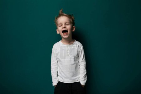 Téléchargez les photos : Un jeune garçon les yeux fermés devant un mur vert vibrant, mettant en valeur un moment de réflexion ou de concentration. Son langage corporel suggère un sentiment de calme et de contemplation. - en image libre de droit