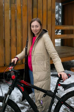 Una mujer está parada junto a una bicicleta en una escena invernal nevada. Ella parece estar preparándose para montar en bicicleta a pesar del clima frío.