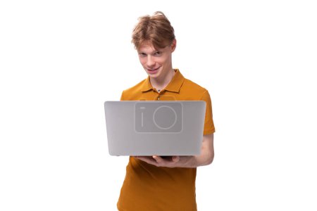 jeune homme roux dans un t-shirt orange étudie en utilisant un ordinateur portable sur un fond blanc avec espace de copie.