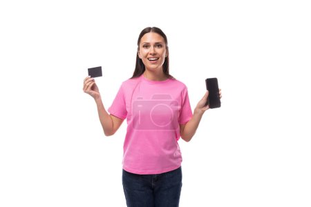 Foto de Joven mujer morena delgada en camiseta básica rosa con tarjeta de crédito maqueta y teléfono inteligente sobre fondo blanco con espacio para copiar. - Imagen libre de derechos