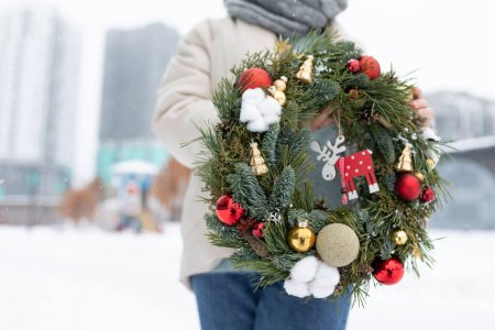 Eine Person steht im Schnee und hält einen Adventskranz in der Hand. Das Individuum wird in Winterkleidung gebündelt, während sie die festliche Dekoration zur Schau stellen.