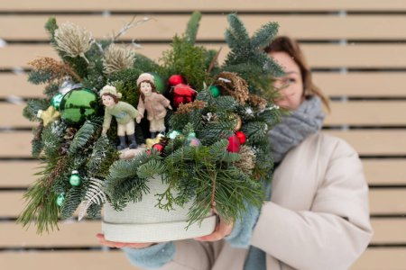 Une femme se tient debout, tenant une plante en pot ornée de décorations de Noël. Elle semble mettre en valeur la plante festive, qui comporte des boules scintillantes, des rubans et des lumières.