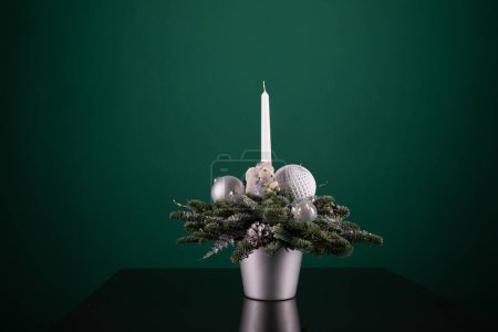 Eine silberne Vase mit einer Vielzahl von Weihnachtsdekorationen wie Kugeln, Bändern und Ornamenten, neben einer weißen Kerze, die hell brennt. Die Szene ist festlich und sorgt für einen Hauch von Feiertagsstimmung