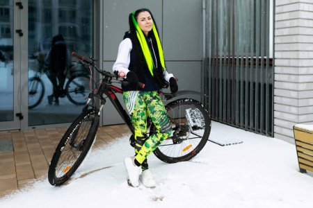 Eine Frau steht neben einem Fahrrad in einer verschneiten Landschaft. Sie trägt warme Kleidung, und es gibt Fußabdrücke im Schnee. Das Fahrrad ist teilweise mit Schnee bedeckt.