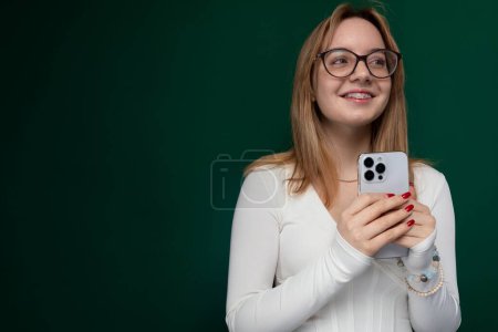 Se ve a una mujer con gafas sosteniendo un teléfono celular en la mano. Parece estar mirando la pantalla atentamente, posiblemente enviando mensajes de texto o navegando. El fondo está borroso, poniendo el foco en el