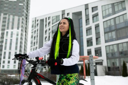 Eine Frau steht neben einem Fahrrad in einer verschneiten Landschaft. Sie trägt warme Winterkleidung und scheint das Fahrrad zu begutachten. Der schneebedeckte Boden und die Bäume bilden eine kalte Kulisse für die