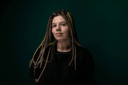 A woman with dreadlocks is standing in front of a solid green background. She is looking directly at the camera with a neutral expression. Her long dreadlocks fall past her shoulders, adding texture
