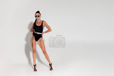 Photo for In a softly lit studio, a mesmerizing woman undresses with grace, unveiling her sleek black lingerie and taut body, while her long ponytail cascades down her back, creating a glamorous composition - Royalty Free Image