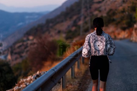 Foto de Vista trasera de una excursionista caminando por un camino de asfalto en las montañas. pantalones cortos negros y chaqueta blanca. Concepto de viaje - Imagen libre de derechos