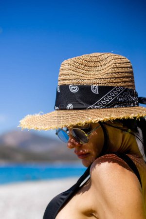 Photo for Back view of young woman in white swimsuit and lovely hat sitting at white sand beach with turquoise sea looking toward the sea - Royalty Free Image