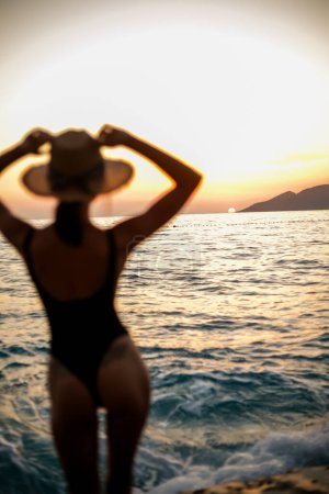 Téléchargez les photos : Avec le coucher du soleil comme toile de fond, une fille tendre dans un élégant maillot de bain noir et chapeau large se tient sereinement près des vagues de la mer - en image libre de droit