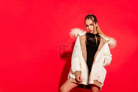 Téléchargez les photos : Une jeune fille respire la confiance alors qu'elle pose en studio sur un fond rouge vif, élégamment ornée d'une veste blanche élégante - en image libre de droit
