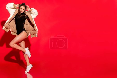 Foto de Sobre un fondo rojo vibrante, una joven emana confianza y encanto en el estudio, su chaqueta blanca de moda agregando un toque de sofisticación a la escena - Imagen libre de derechos