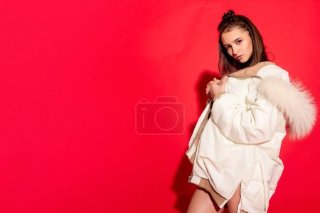 Foto de Encanto exuberante y elegancia, una joven toma una pose en el estudio contra un fondo rojo audaz, su chaqueta blanca de moda completando el look - Imagen libre de derechos