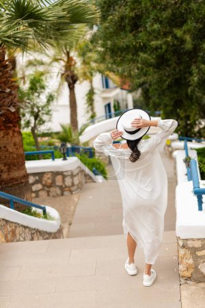 Foto de Paraíso tropical: Una chica con un encantador vestido blanco y sombrero de paja deambula entre casas blancas y palmeras, añadiendo un toque de fantasía a la escena - Imagen libre de derechos