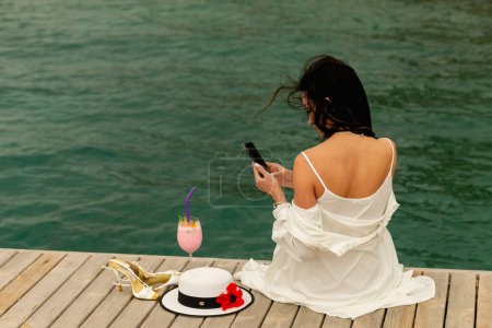 Foto de Elegancia bañada por el sol: Una niña con un vestido blanco radiante y un sombrero de paja disfruta del paisaje costero de casas blancas y palmeras, exudando encanto tropical - Imagen libre de derechos